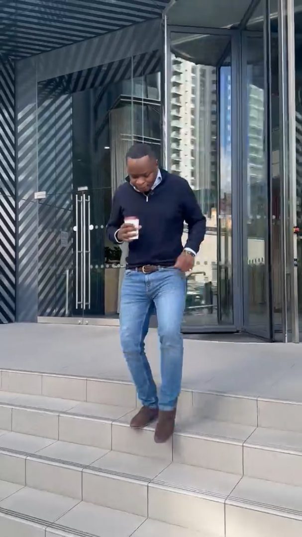 Nos › man in a navy sweater and jeans stepping down stairs outside a glass storefront, holding a cup of coffee.