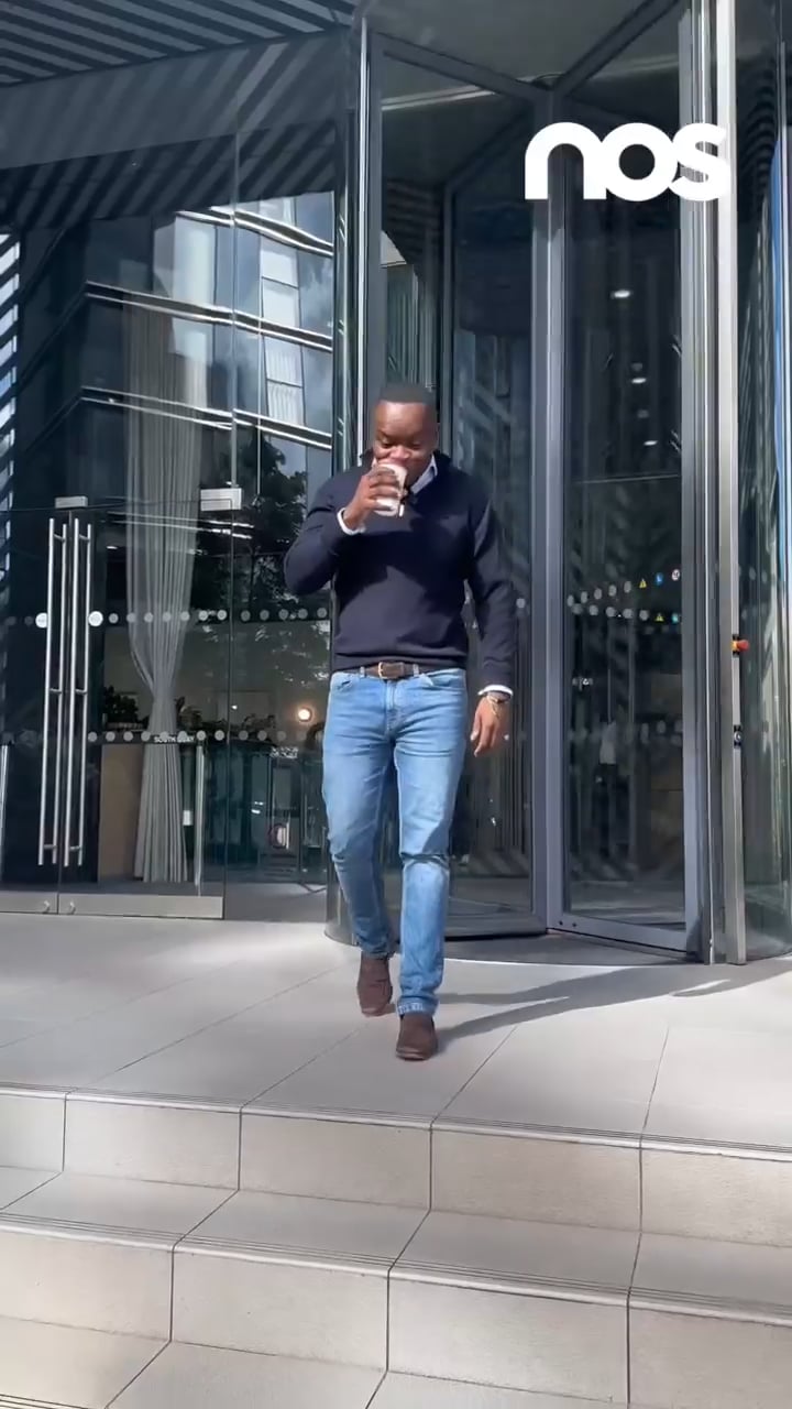 Man in a navy sweater and jeans walking down tiled steps while sipping a coffee outside a glass building with a NOS logo above the door.