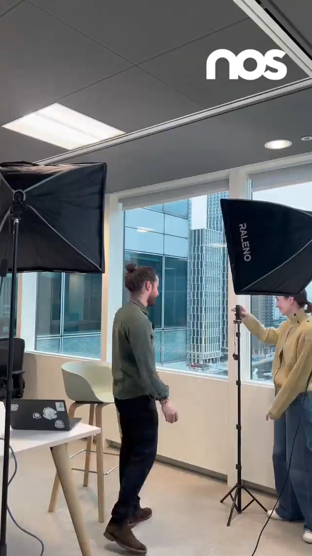 Two people in a bright office studio adjusting large photography lights for a shoot, with a laptop on a nearby table and windows in the background
