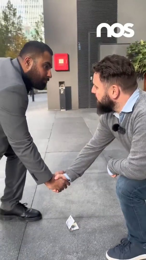 Nos › two men in business attire shake hands on a sidewalk outside a modern building, one leaning forward as they greet, with a red fire alarm and logo in the background.
