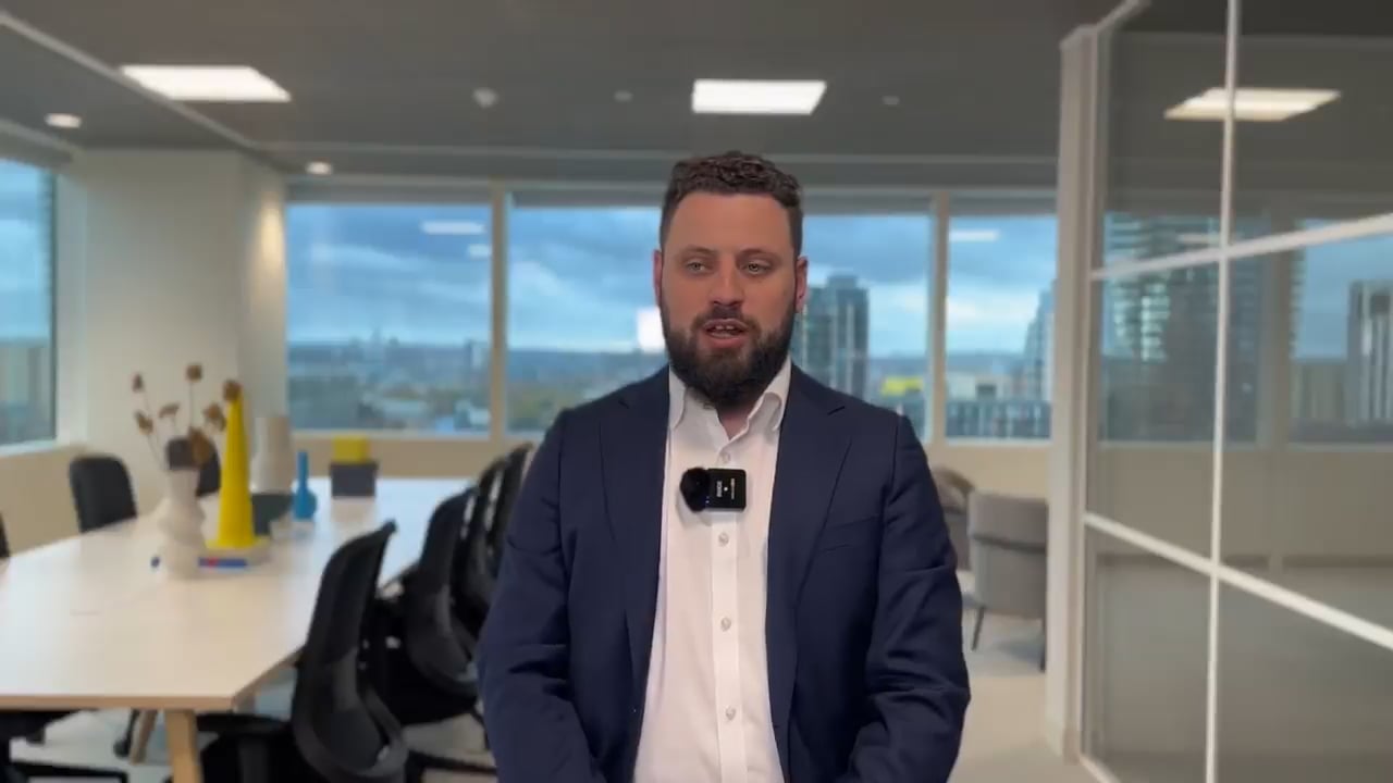 Nos › man in a navy suit and white shirt seated in a modern office, wearing a lavalier mic and speaking to camera with cityscape visible outside the windows.