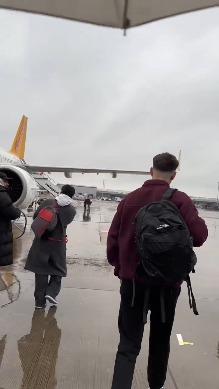 Passengers with backpacks walk on a wet tarmac toward a white airplane with an orange tail under a gray, cloudy sky.