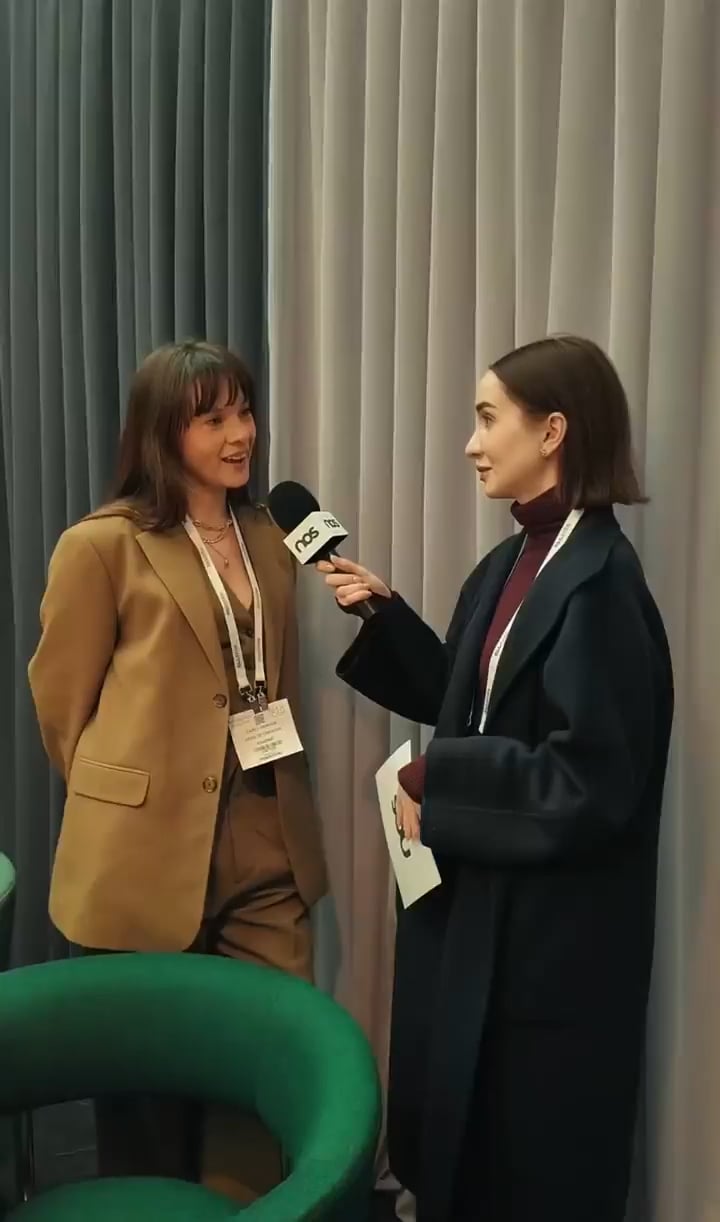 Two women in a hallway interview: the interviewer holds a NOS microphone toward a woman in a tan suit with a name badge.
