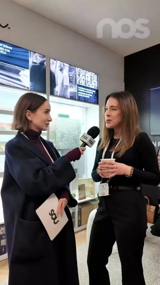 Nos › two women in a hallway interview: a reporter holds a nos microphone toward a woman in black, both smiling. Nos logo visible above.