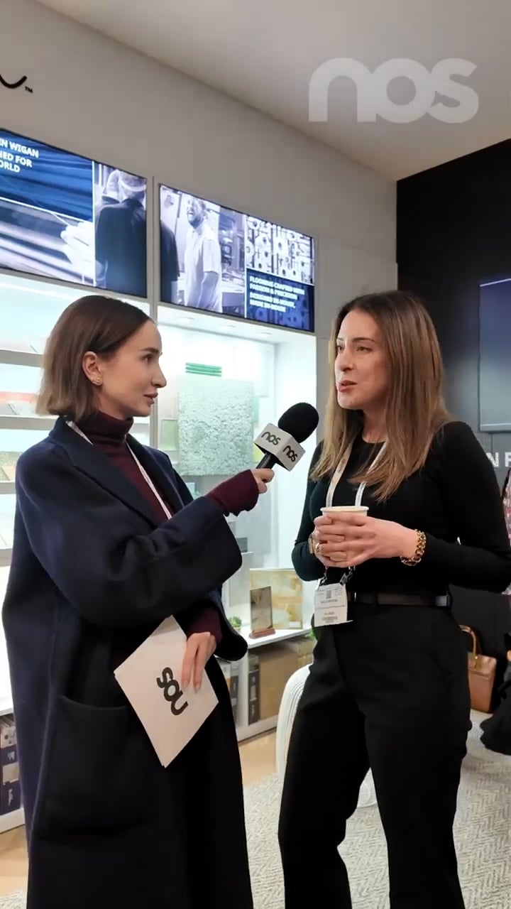 Two women interview each other indoors: a reporter holds a NOS microphone toward a speaker who holds a cup, with display cases in the background.