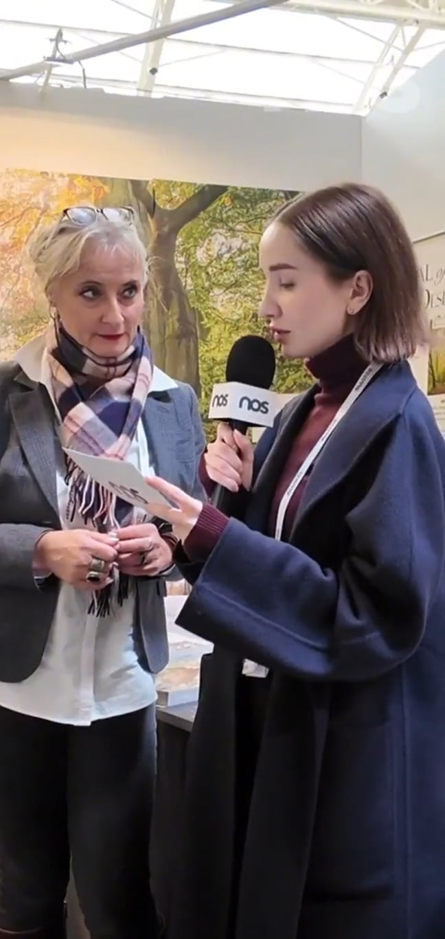 Interviewer with NOS microphone filming a woman who holds a paper at an indoor event, forest mural backdrop behind them.