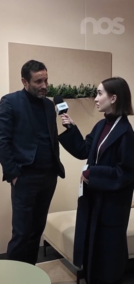 Nos › man in a dark suit being interviewed by a woman holding a nos microphone in a neutral indoor setting.
