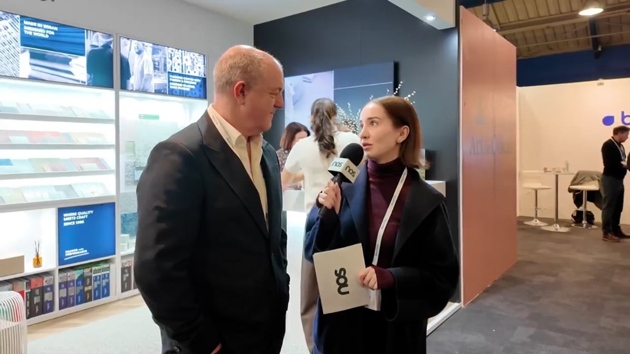 Nos › female reporter interviews a man at a trade show booth; she holds a folder with a logo and a microphone close to his face.