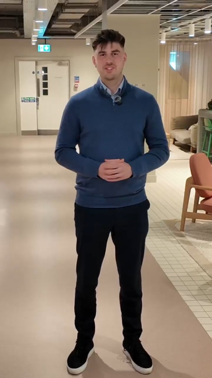 Young man in a blue sweater with a lavalier mic, standing in a modern office lobby.