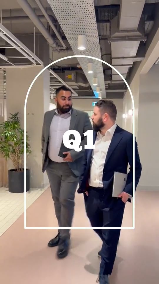 Nos › two businessmen in suits walk together down a modern office hallway, one carrying a tablet and the other a laptop, with a bold 'q1' overlay.