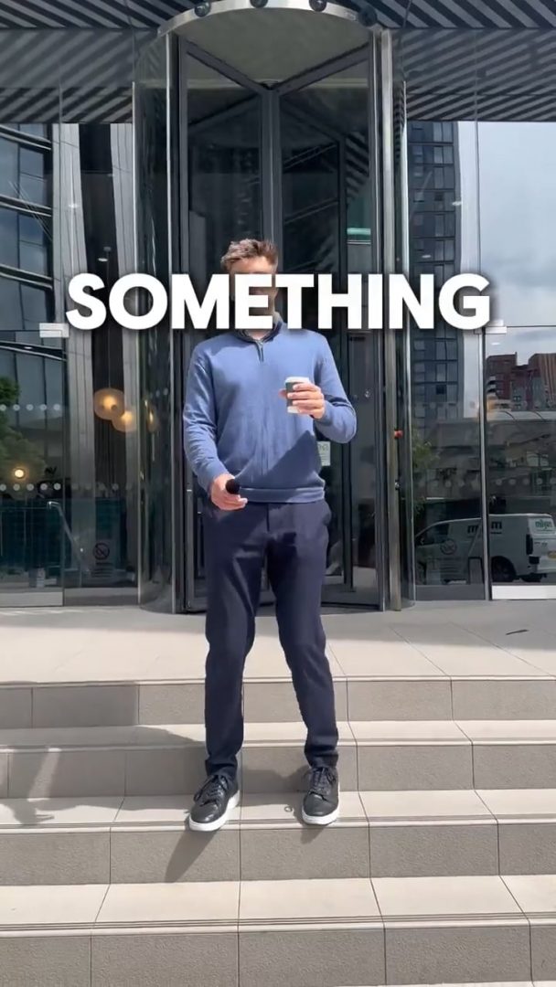 Nos › man in a blue pullover stands on steps outside a glass-front building, holding a coffee cup and a smartphone.