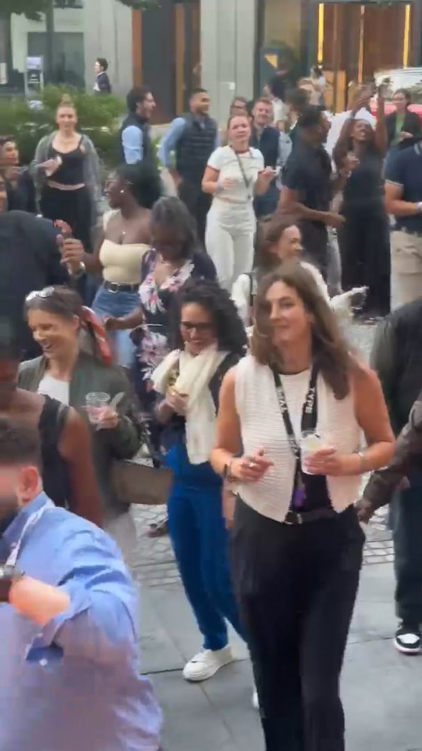 Nos › crowd of people walking and socializing outdoors at an event, many holding cups.