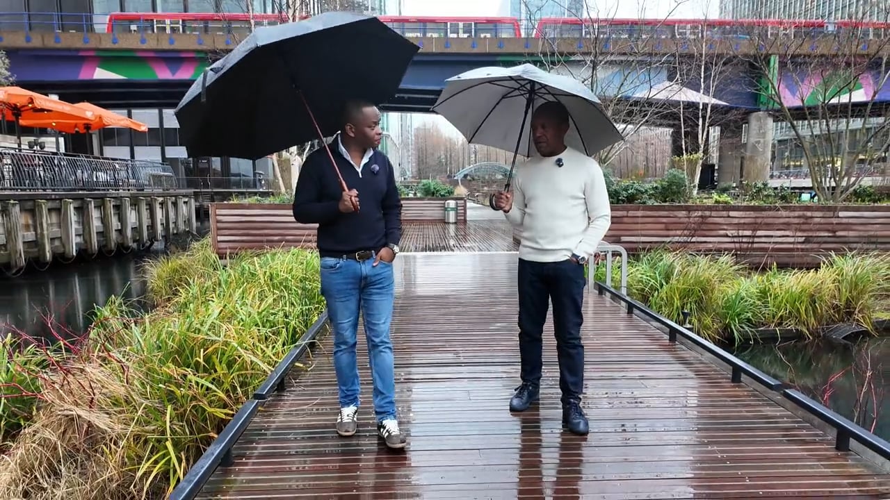 Nos › two men stand on a wet wooden boardwalk, each holding an umbrella and chatting on a rainy day in an urban park.