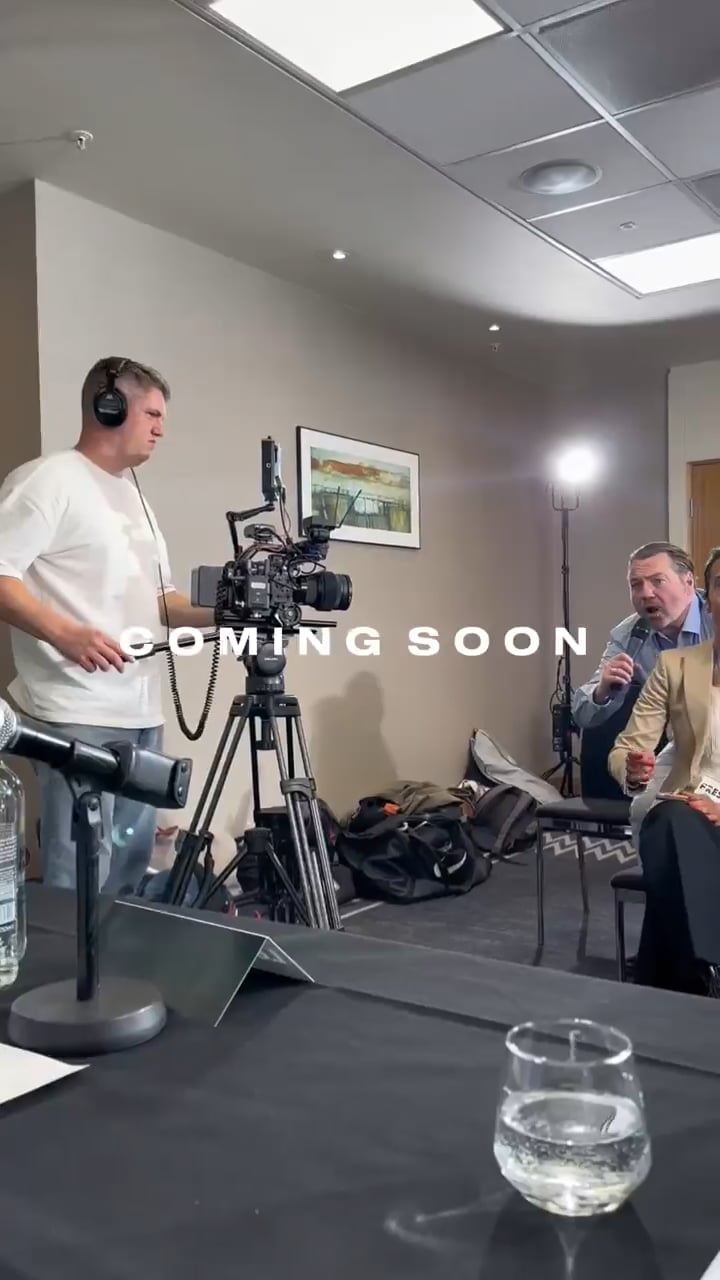 Camera operator filming a panel discussion in a studio, with a tripod-mounted camera and a spread-water bottle on the table; 'COMING SOON' text overlay appears across the frame.