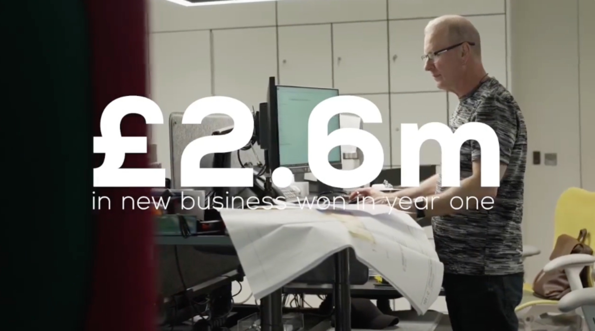 Man standing at a desk in an office, with large white text overlay reading “£2.6m in new business won in year one.”
